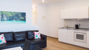 Ett modernt vardagsrum med en svart lädersoffa, ett litet bord och en färgglad kudde. Intill vardagsrummet finns ett kompakt kök med vita skåp och en inbyggd ugn. På väggen hänger en inramad bild som föreställer ett tropiskt landskap.