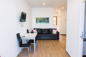 A compact living space with a dining area, a black sofa with colorful cushions, a flat-screen TV mounted on the wall, and a large window providing a view of the outdoors.
