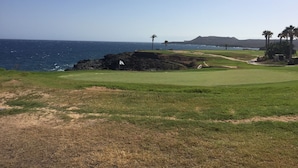 A coastal golf course with a flagstick on the green, a bunker, and a distant mountain.