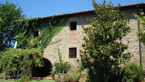 A stone building with a tiled roof, a large tree in front, and a grassy area.