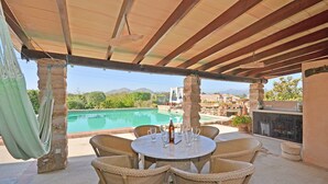 Un espacio cubierto con mesas y sillas de mimbre, con vistas a una piscina y un paisaje montañoso.