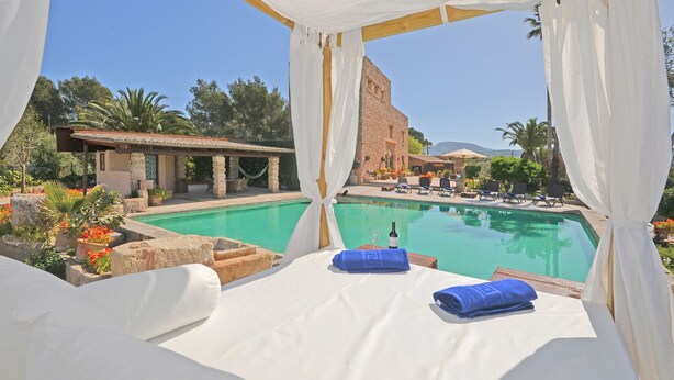Una cama junto a la piscina con ropa blanca y toallas azules, con vistas a una piscina de aguas cristalinas, rodeada de exuberante vegetación y un edificio con tejado de azulejos.