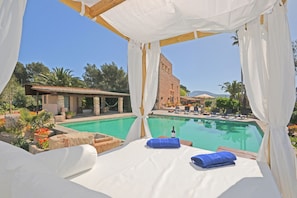 A poolside bed with white linens and blue towels, overlooking a swimming pool with clear blue water, surrounded by lush greenery and a building with a tiled roof. - Torreon - Countryside house with pool 150 meters from the beach in Alcúdia (Alcúdia)