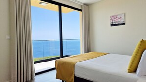 A hotel room with a large window offering a view of the sea, a bed with a yellow throw, and a painting on the wall. - Eden Sun - Luxury Villa w/ocean View by LovelyStay (Prazeres)
