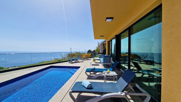 A modern outdoor pool area with lounge chairs and a view of the sea. - Eden Sun - Luxury Villa w/ocean View by LovelyStay (Prazeres)