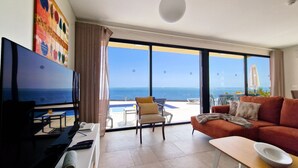 A modern living room with a TV, a sofa, a coffee table, and a view of the ocean through large windows. - Eden Sun - Luxury Villa w/ocean View by LovelyStay (Prazeres)