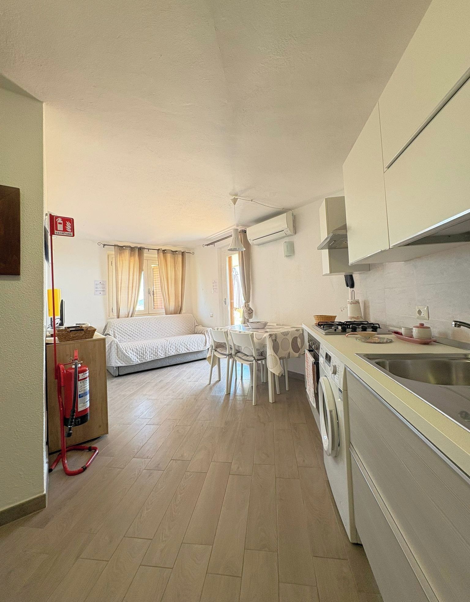 A compact kitchen with a washing machine, a dining table with chairs, and a bed in the background.