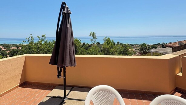A terrace with a table covered by a patterned tablecloth, a chair, and an umbrella. - New valmar Two-room apartment 56 - Via del Lentischio (Costa Rei)