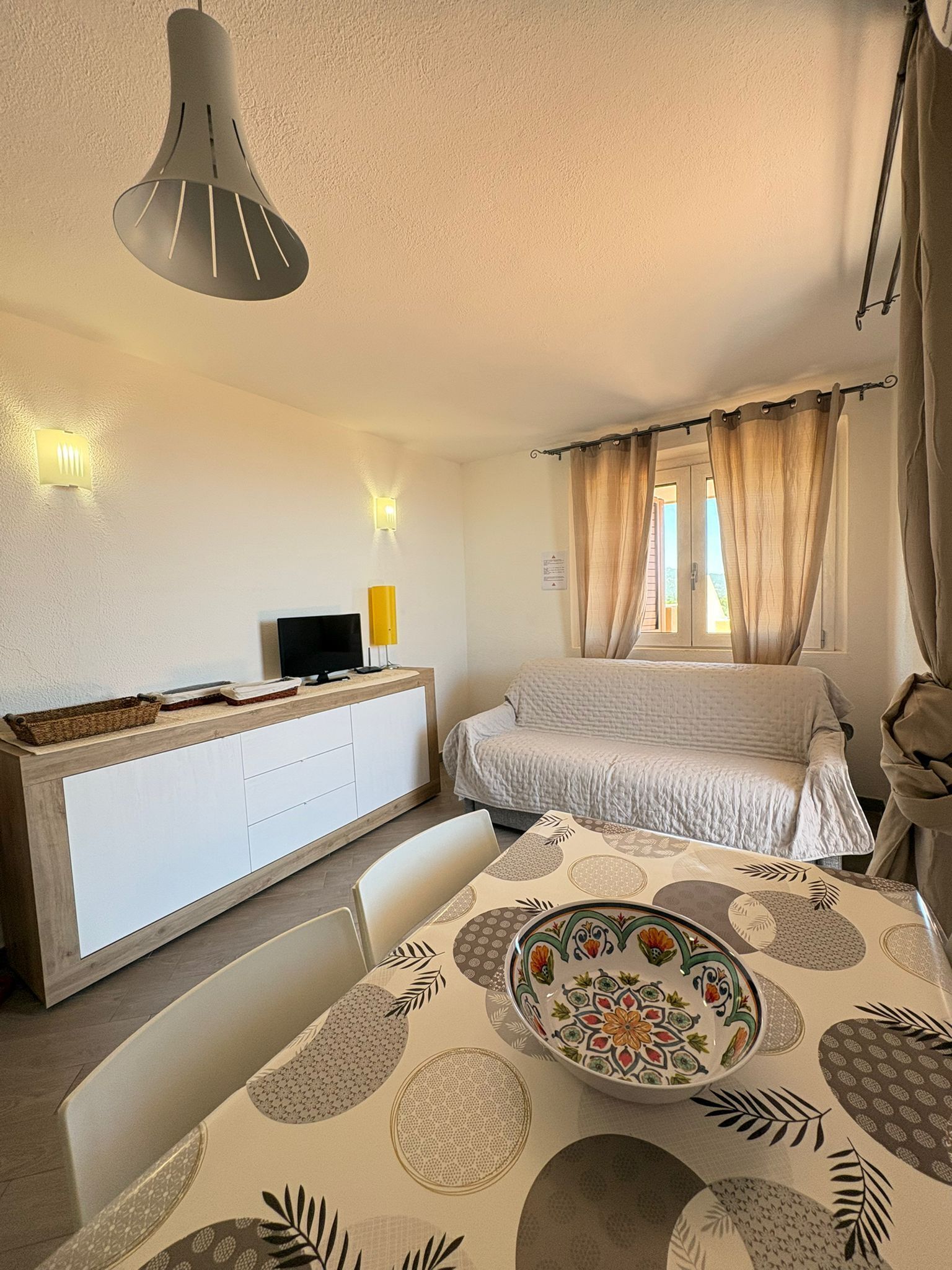 A bedroom with a bed, a bedside table, a desk with a computer, a window with curtains, and a dining area with a table and chairs.