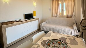 A bedroom with a bed, a bedside table, a desk with a computer, a window with curtains, and a dining area with a table and chairs. - New valmar Two-room apartment 56 - Via del Lentischio (Costa Rei)