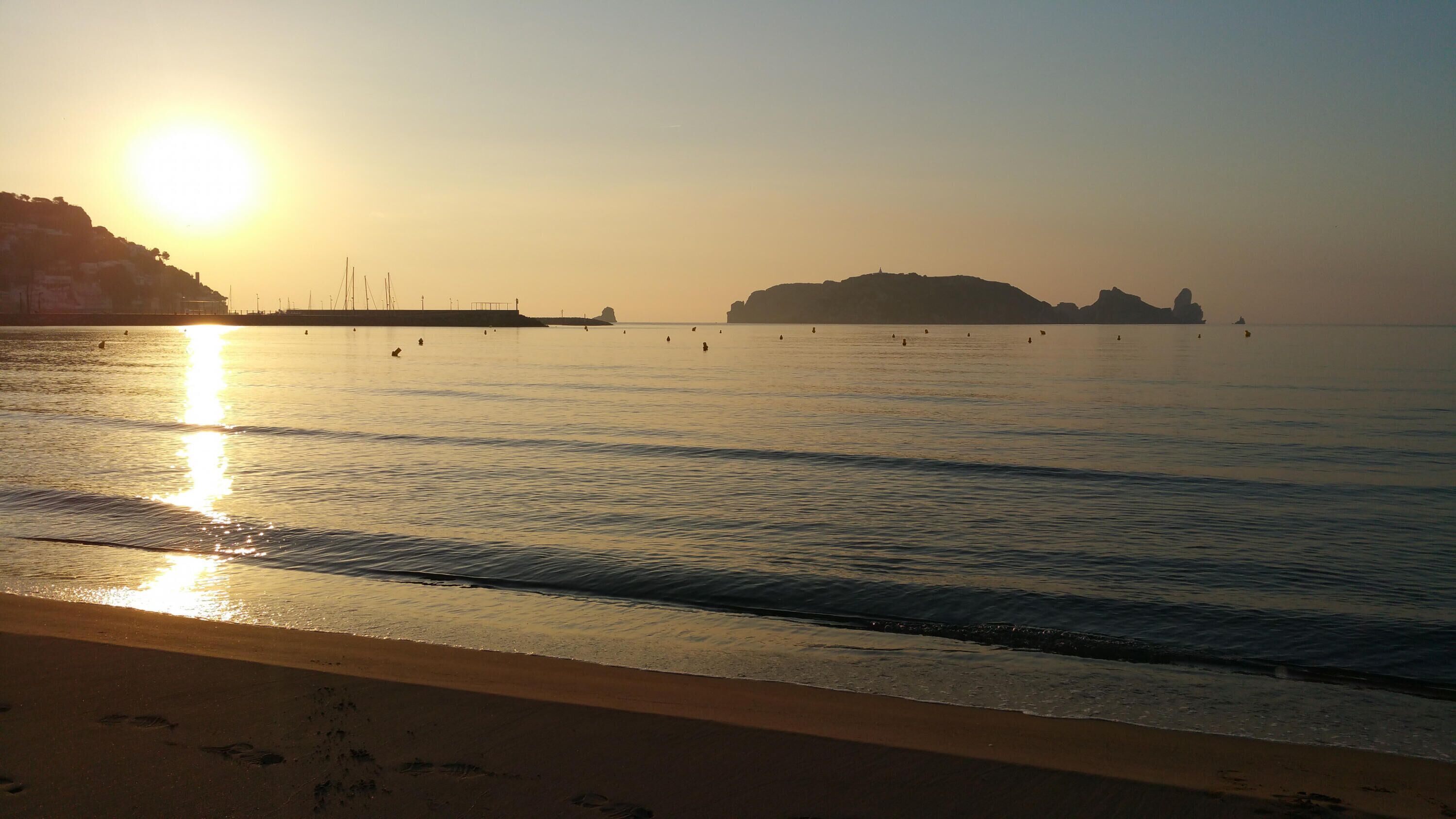 A beach with calm waters reflecting the sun, a sandy shore, and distant rocky formations.