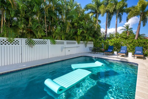 A swimming pool with two blue floating loungers, a white fence, and palm trees in the background.