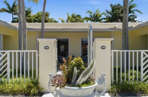 A yellow building with a gated entrance, palm trees, and a white decorative planter with succulents.