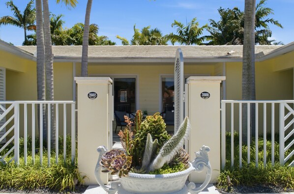 A yellow building with a gated entrance, palm trees, and a white decorative planter with succulents.