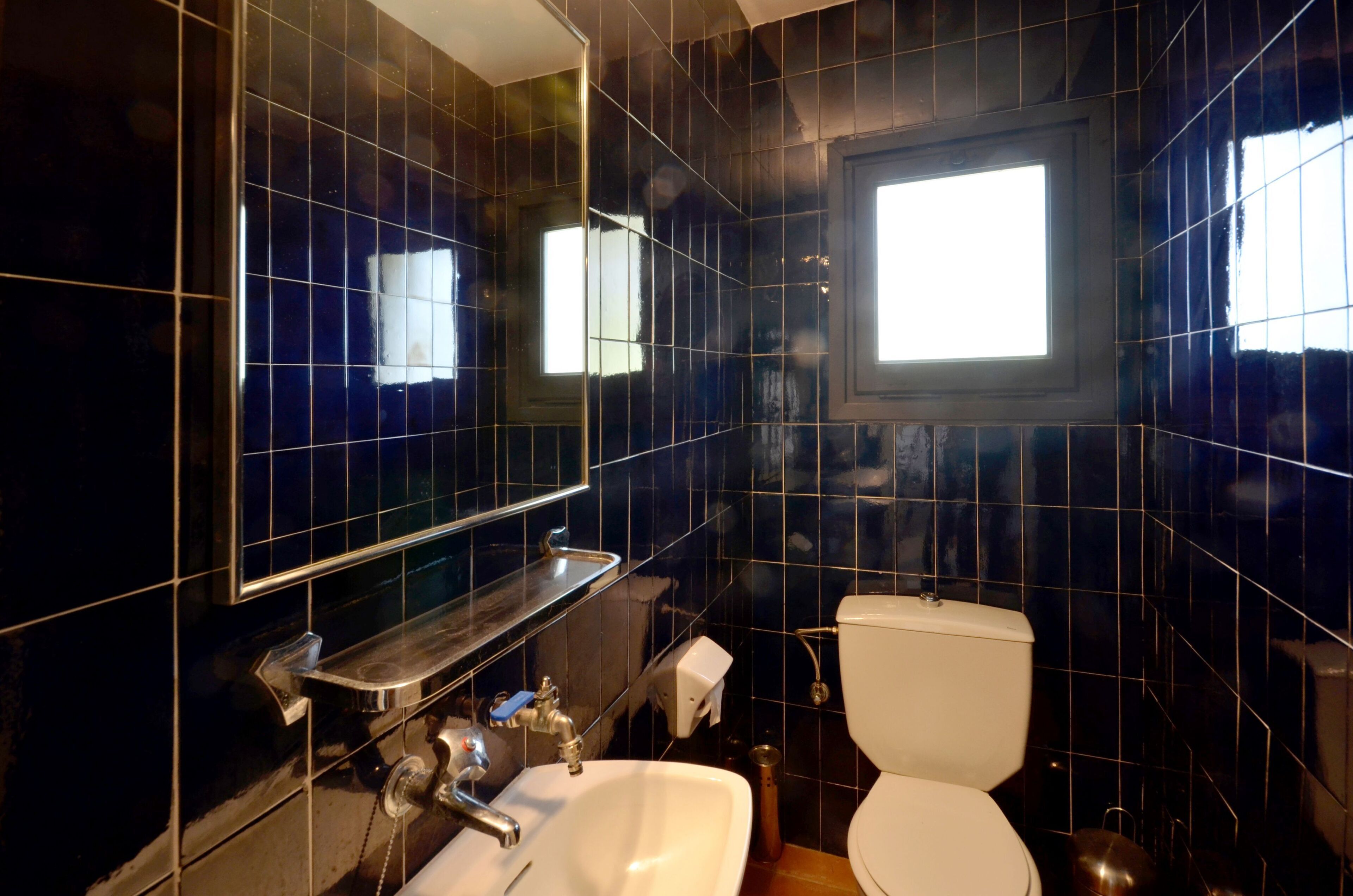 Une salle de bain avec des carreaux bleu foncé, un lavabo blanc, un miroir et une fenêtre.