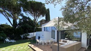 Une terrasse extérieure moderne, avec une table et des chaises blanches, un canapé en osier et une pergola offrant de l’ombre.