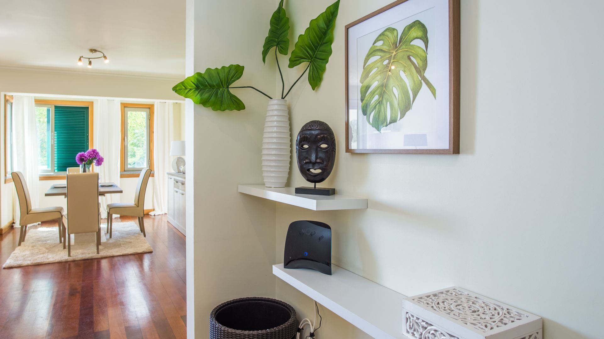 A modern living room with a dining area, featuring a large plant, a framed artwork, and a decorative mask on a shelf.