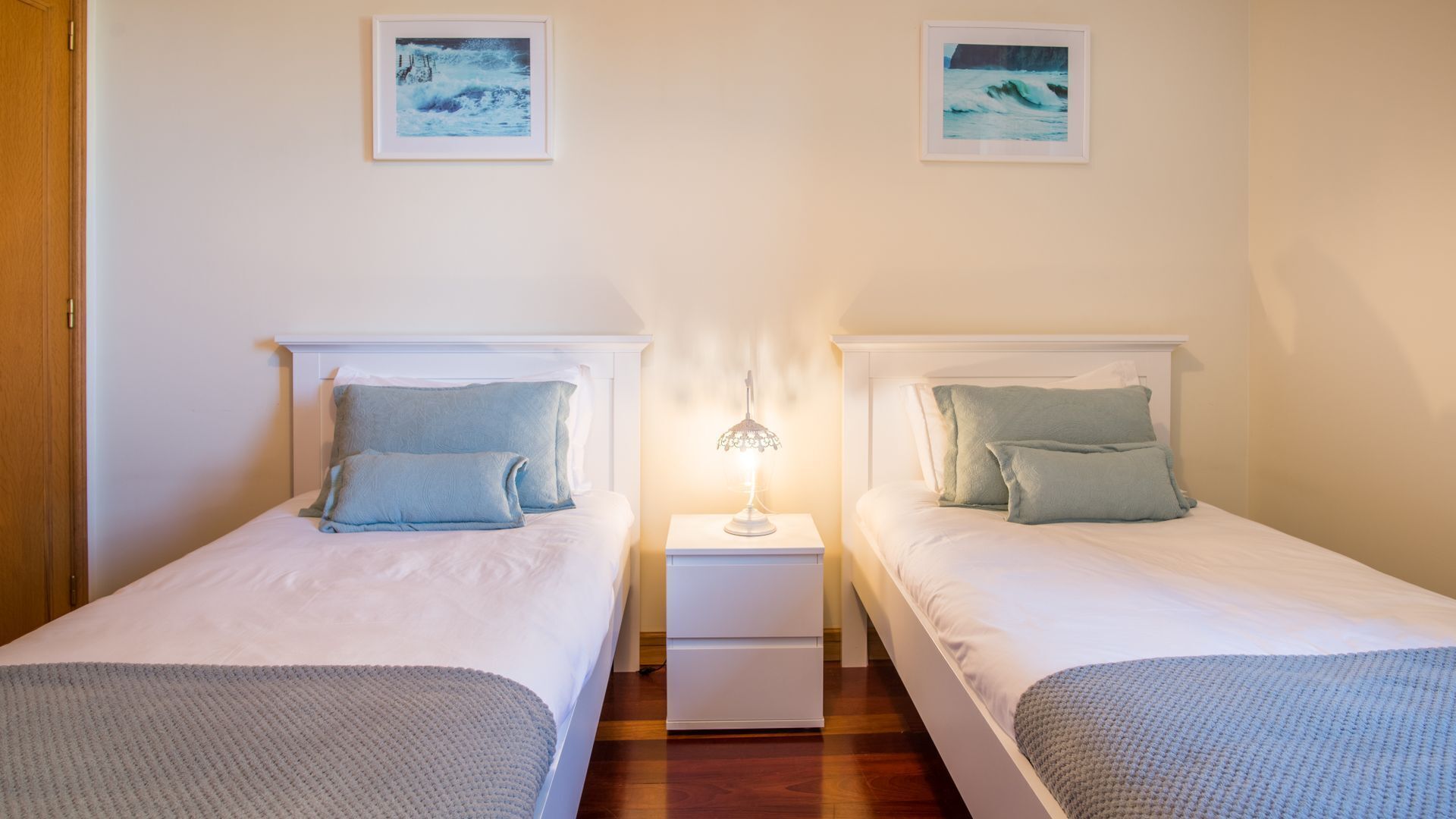 Ein Zimmer mit zwei Einzelbetten, einem Nachttisch mit Lampe sowie zwei gerahmten Bildern an der Wand.