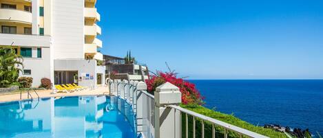 Piscina de hotel com vista para o oceano, cercada por vegetação e um edifício branco.