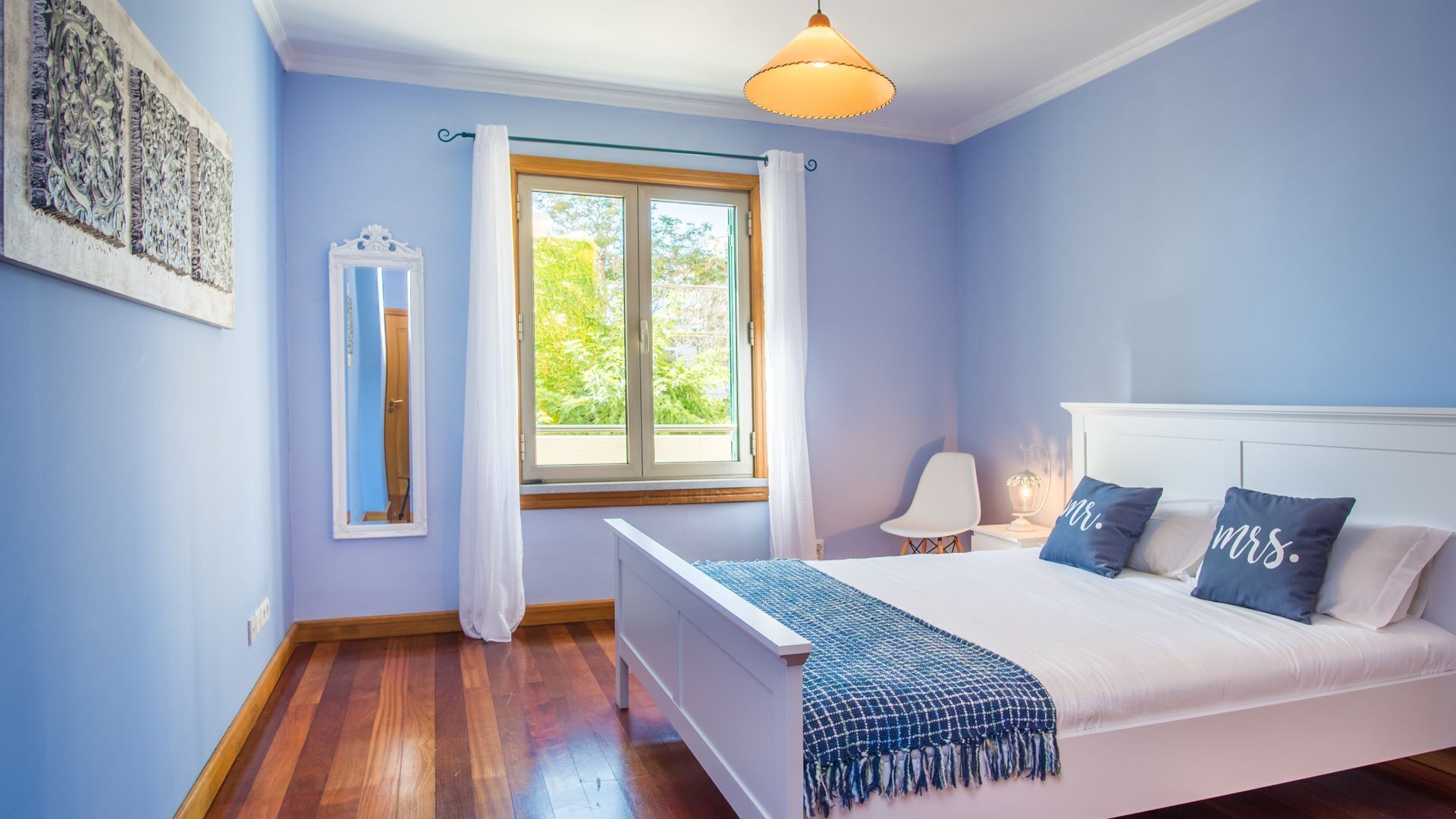 A bedroom with a bed, bedside lamps, a window with curtains, a mirror, and a wall-mounted artwork.