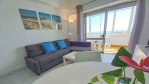 Een woonkamer met een grijze bank, een witte tafel en een potplant. Drie schilderijen hangen aan de muur en er is uitzicht op het buitenlands terrein door een glazen deur.