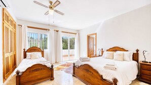 A spacious bedroom with two wooden beds, a ceiling fan, and large windows with curtains.