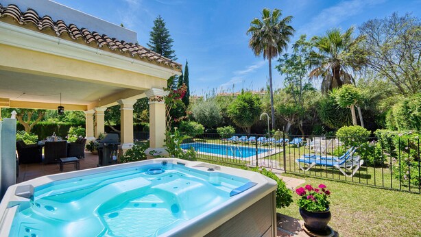 A hot tub in a backyard with a pool, patio furniture, and lush greenery.