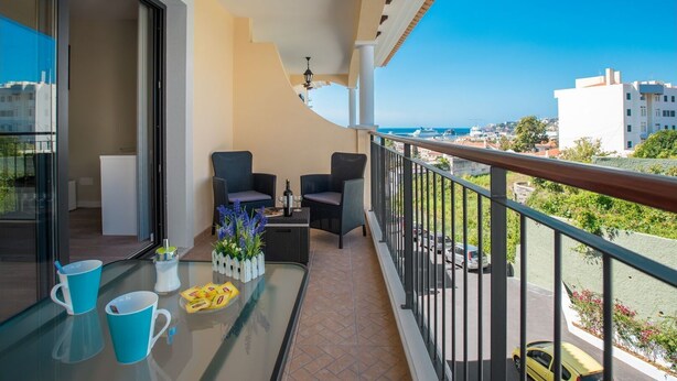 Ein Balkon mit zwei Korbstühlen, einem Glastisch mit Blumenvase und Blick auf das Meer und Gebäude.