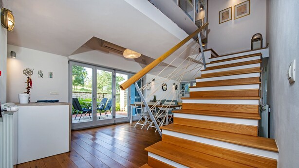 Ein modernes Interieur mit einer Holztreppe, einem Essbereich mit Tisch und Stühlen und Ausblick ins Freie durch große Glasüren.
