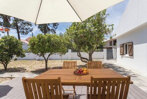 An outdoor patio with a wooden table and chairs, surrounded by trees and a white building.