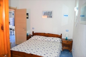 Um quarto com cama de madeira, mesa de cabeceira com abajur, quadro na parede e porta que dá para outro cômodo.