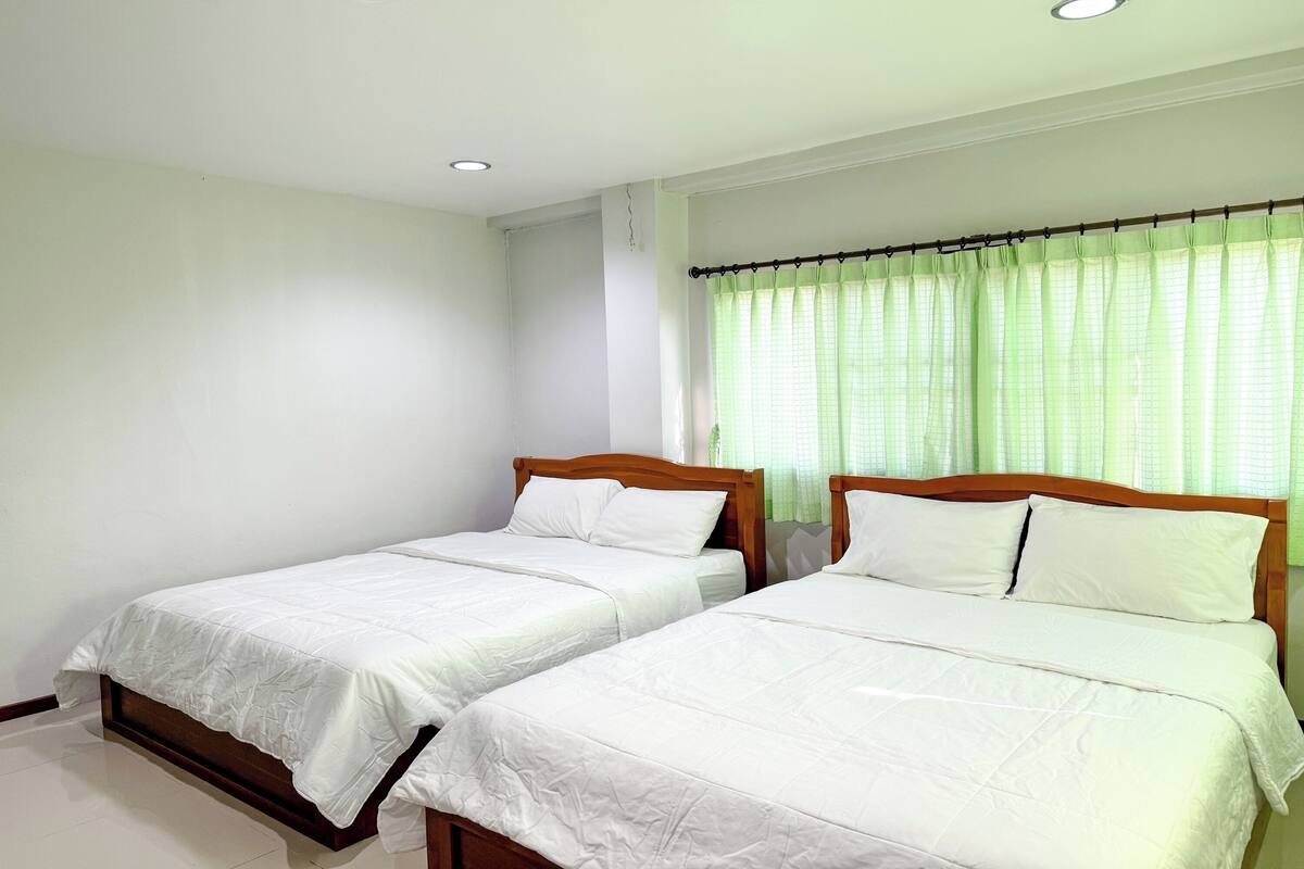A room with two single beds, white bedding, wooden headboards, a window with green curtains, and a white wall.
