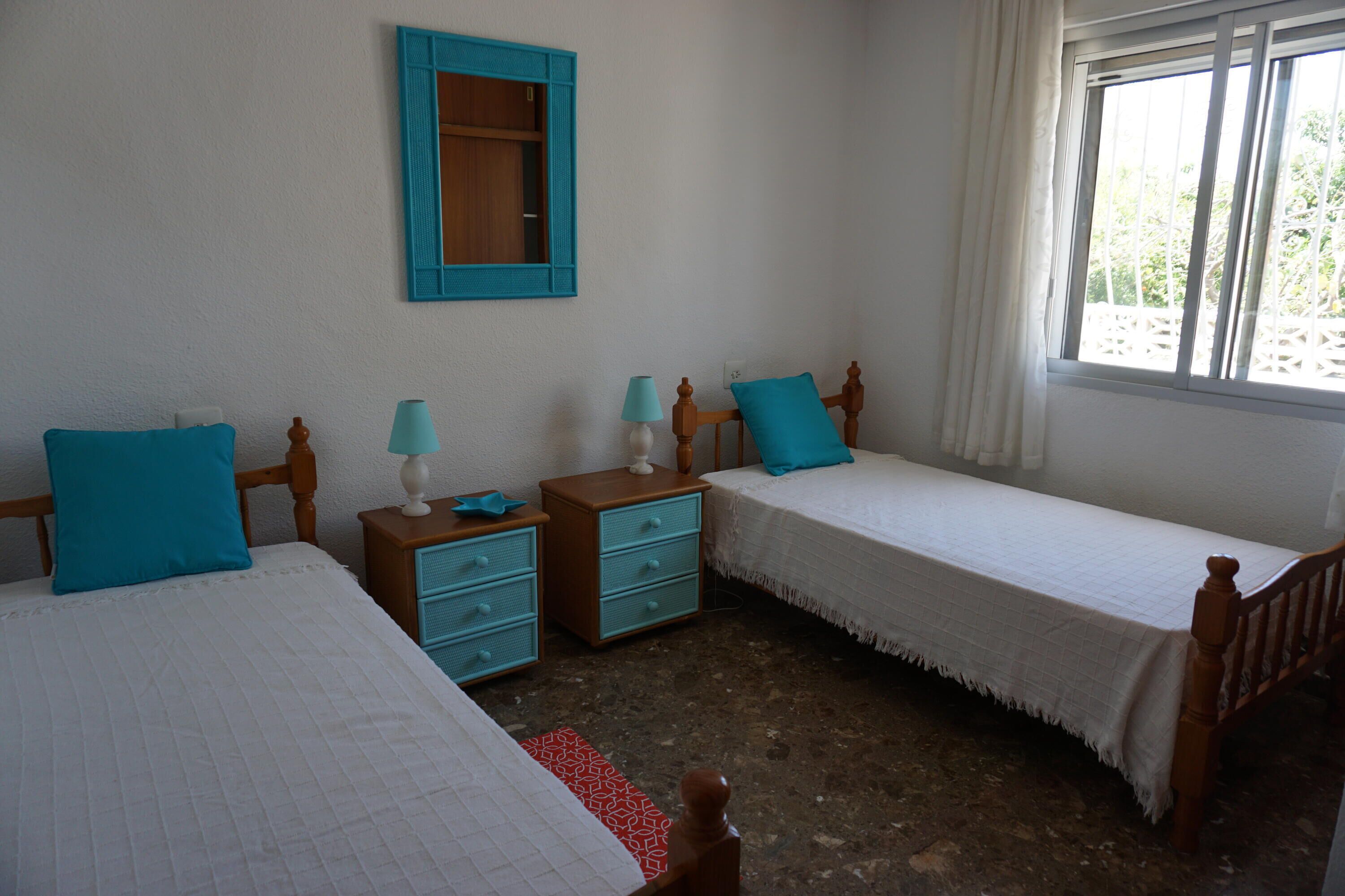 Een slaapkamer met twee eenpersoonsbedden, een nachtkastje met lades, een spiegel en een raam met gordijnen.