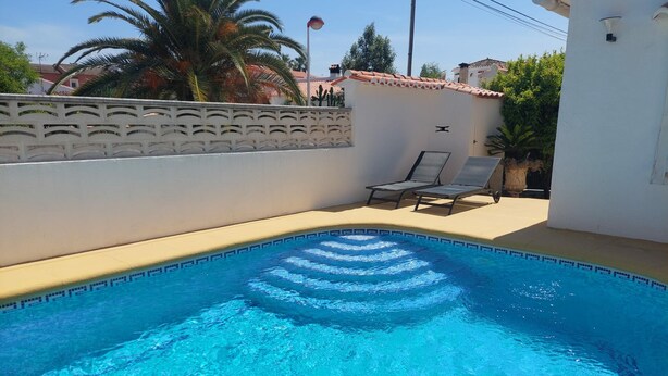 Una piscina con borde de azulejos, dos sillas de descanso y una pared blanca con diseños decorativos.