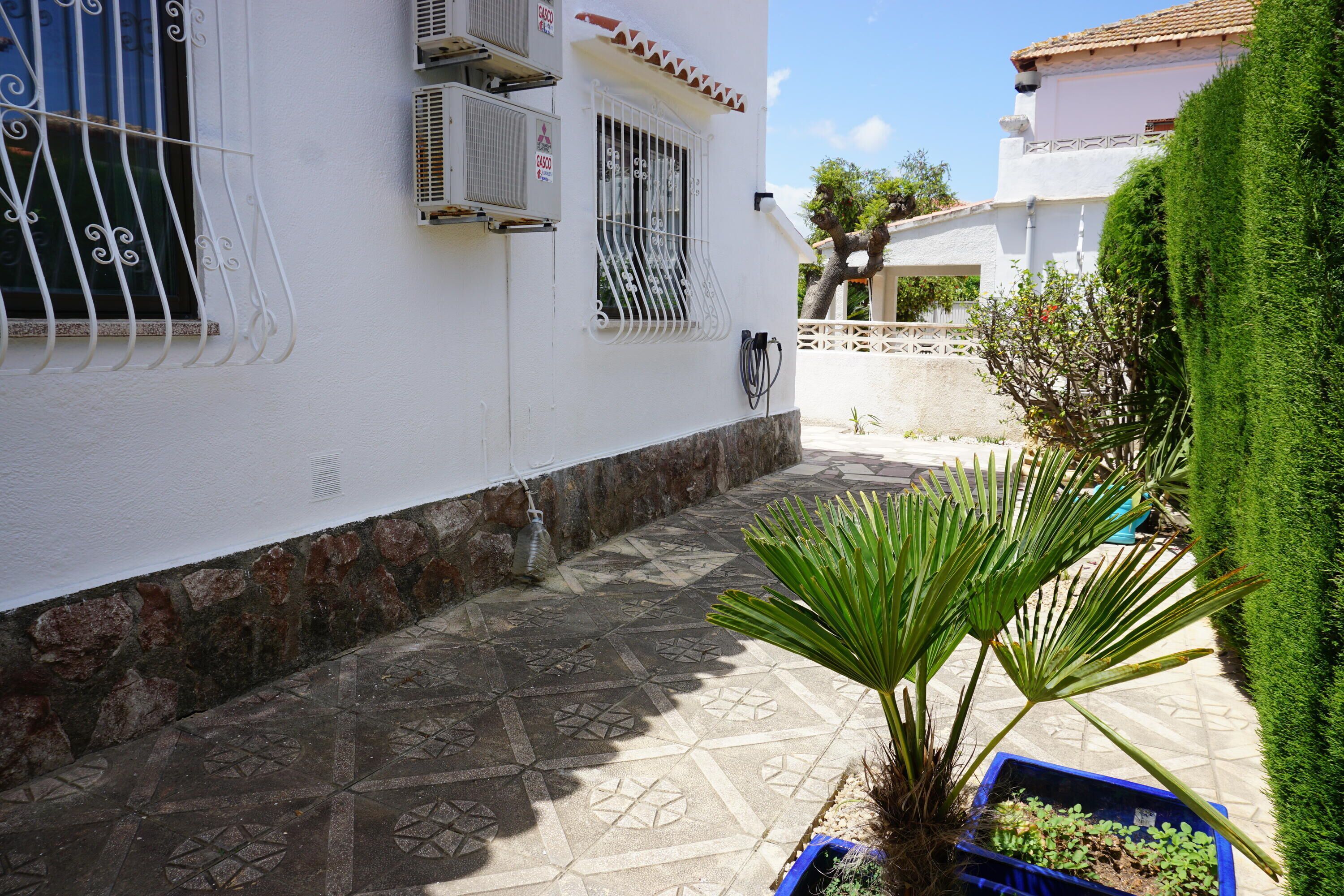 Une cour avec un sol carrelé à motifs, un pot de fleurs et un bâtiment blanc doté de fenêtres grillagées et de climatiseurs.