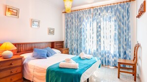 Une chambre à coucher avec un lit, une table de chevet en bois, une lampe, une chaise et une grande fenêtre avec des rideaux bleus.
