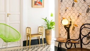 A room with a patterned floor, a green chair, a wooden chair, a table with a lamp, and a wall with a toucan design. - Genteel Home Romanilla (Granada)