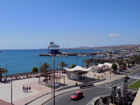 Un paisaje urbano costero con un gran crucero amarrado en un muelle, un puerto deportivo con barcos y un paseo peatonal con palmeras.