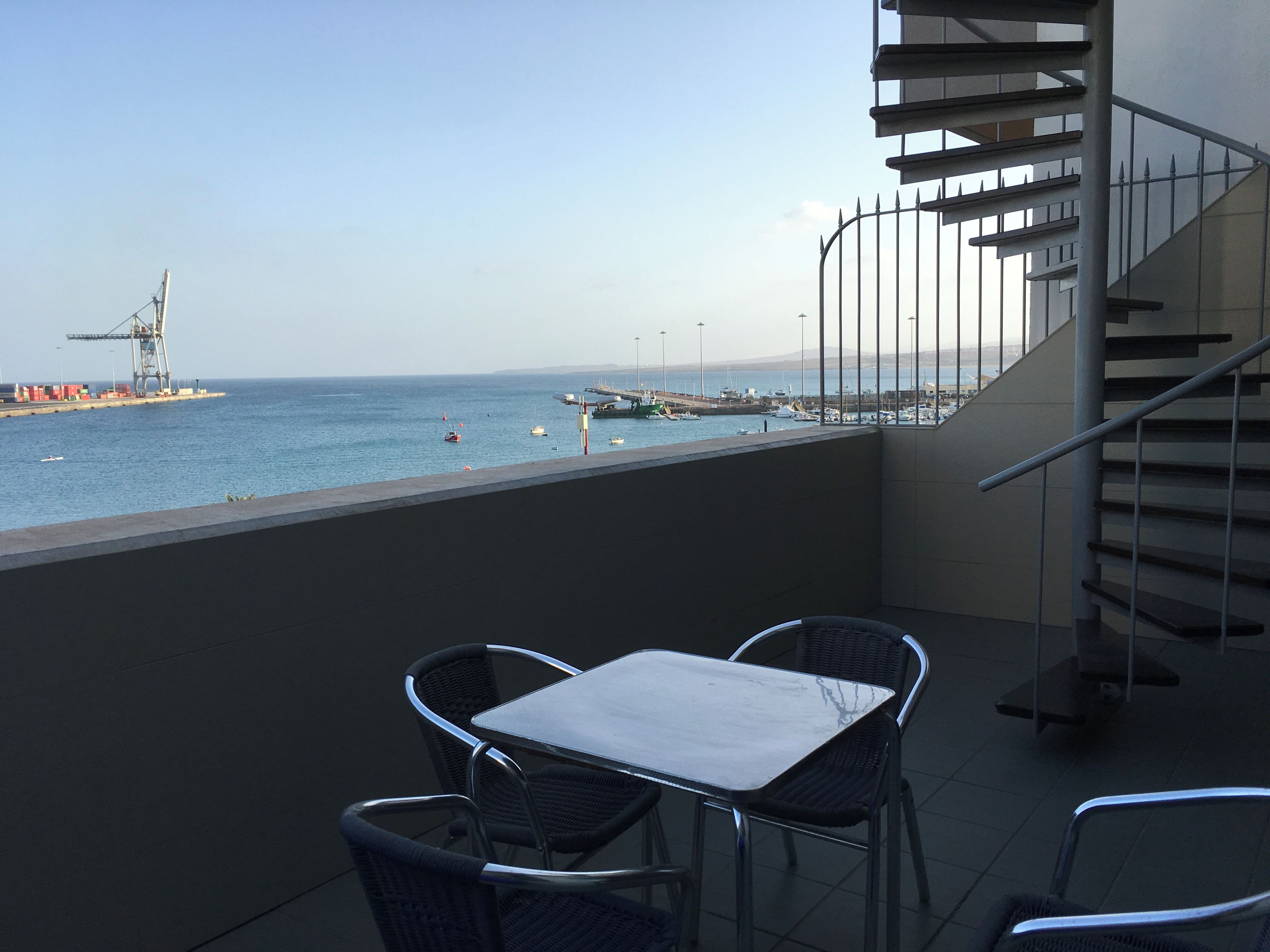 Un balcón con mesa y sillas, con vista a una marina con barcos y un barco a lo lejos.