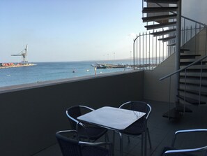 Un balcón con mesa y sillas, con vista a una marina con barcos y un barco a lo lejos.