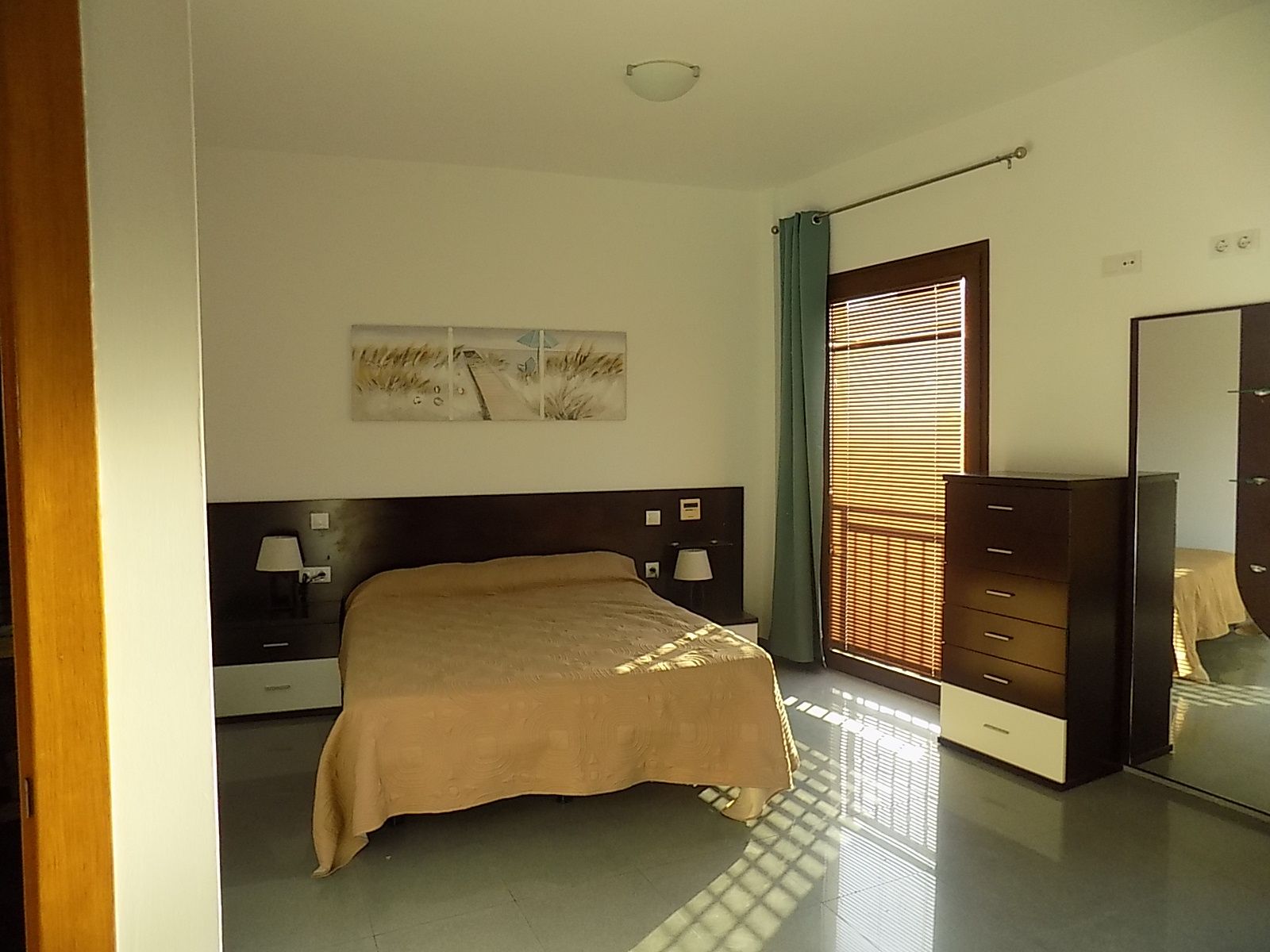 Een slaapkamer met een bed, nachtlampjes, een dressoir, een spiegel en een raam met jaloezieën.