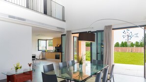 Un espace repas moderne avec une table et des chaises en verre, un banc rouge et une vue sur un jardin et un moulin à vent à l’extérieur.