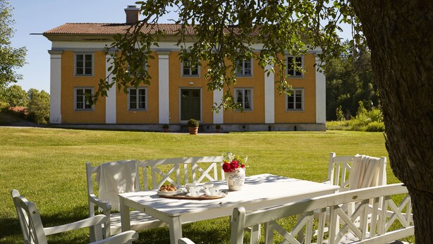 A white outdoor dining set with a table and chairs, a potted plant, and a plate of food on a lawn in front of a yellow building with a green door.