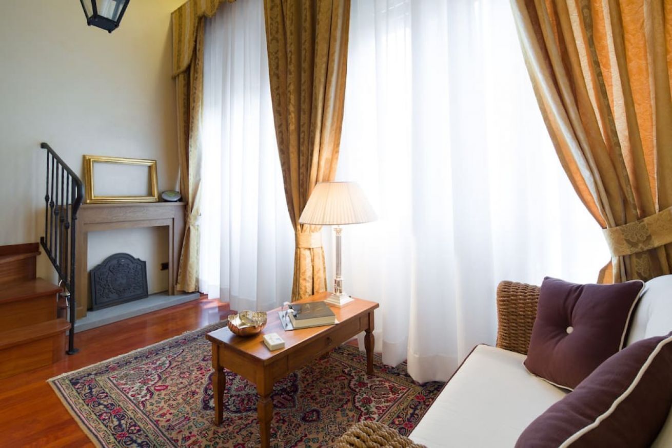 A room with a fireplace, a wooden table with a lamp, a sofa, and a patterned rug.