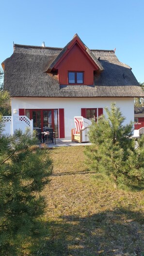 A house with a red roof, white walls, and a thatched roof. There is a patio with outdoor furniture and a small tree in front.
