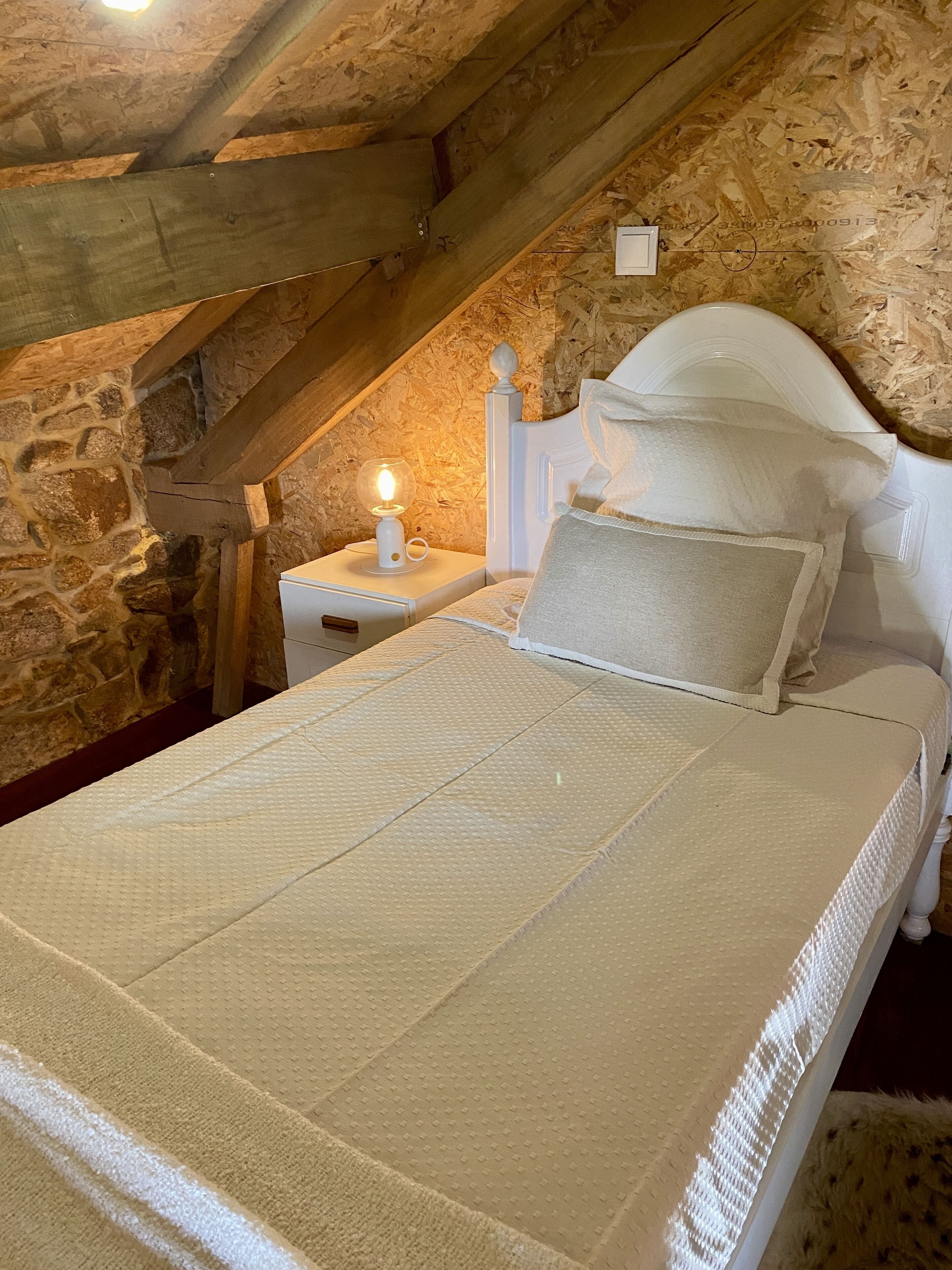 A bedroom with a stone wall, a wooden ceiling, a white bed with a textured cover, and a nightstand with a lamp.