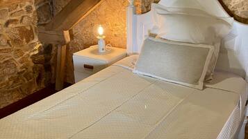 A bedroom with a stone wall, a wooden ceiling, a white bed with a textured cover, and a nightstand with a lamp.