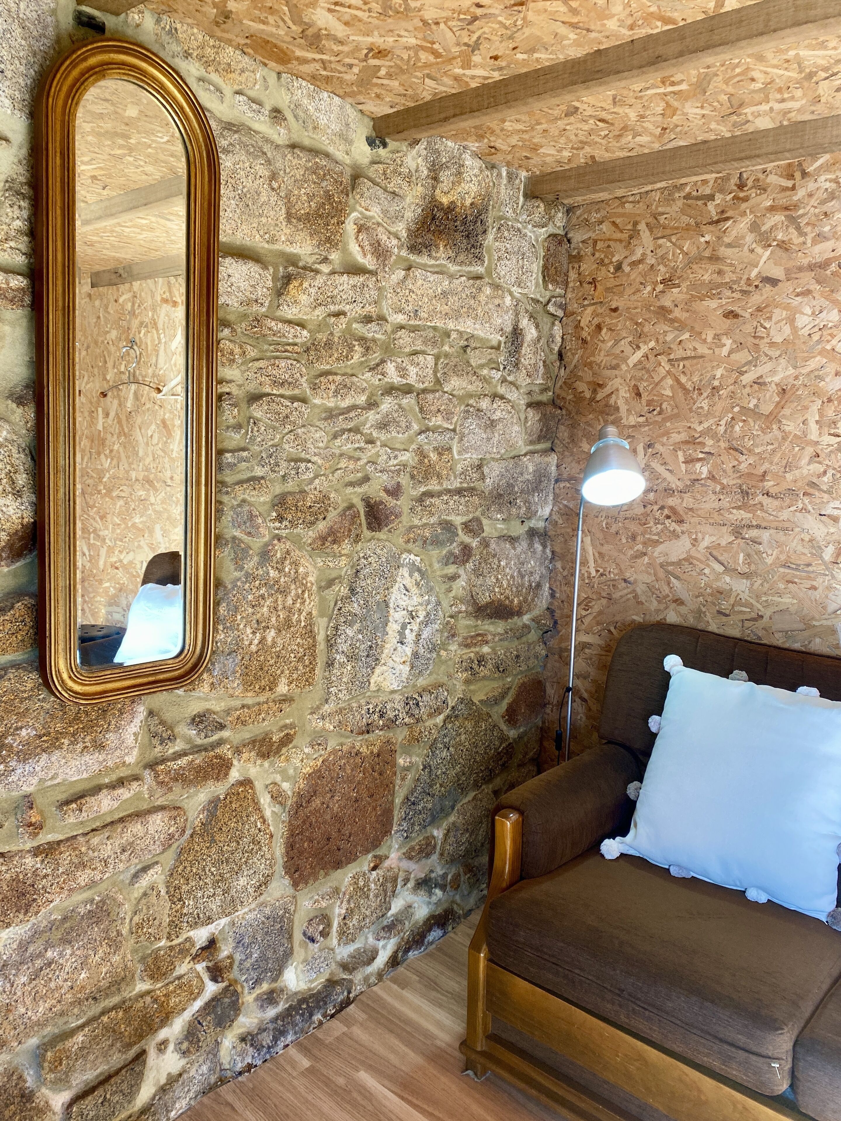 Una stanza con una parete in pietra, uno specchio con cornice in legno, un divano marrone con un cuscino bianco e una lampada da terra.