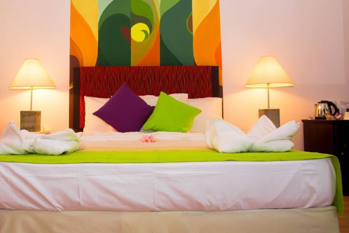 A hotel room with a bed, two bedside lamps, a red chair, and a colorful abstract painting on the wall.