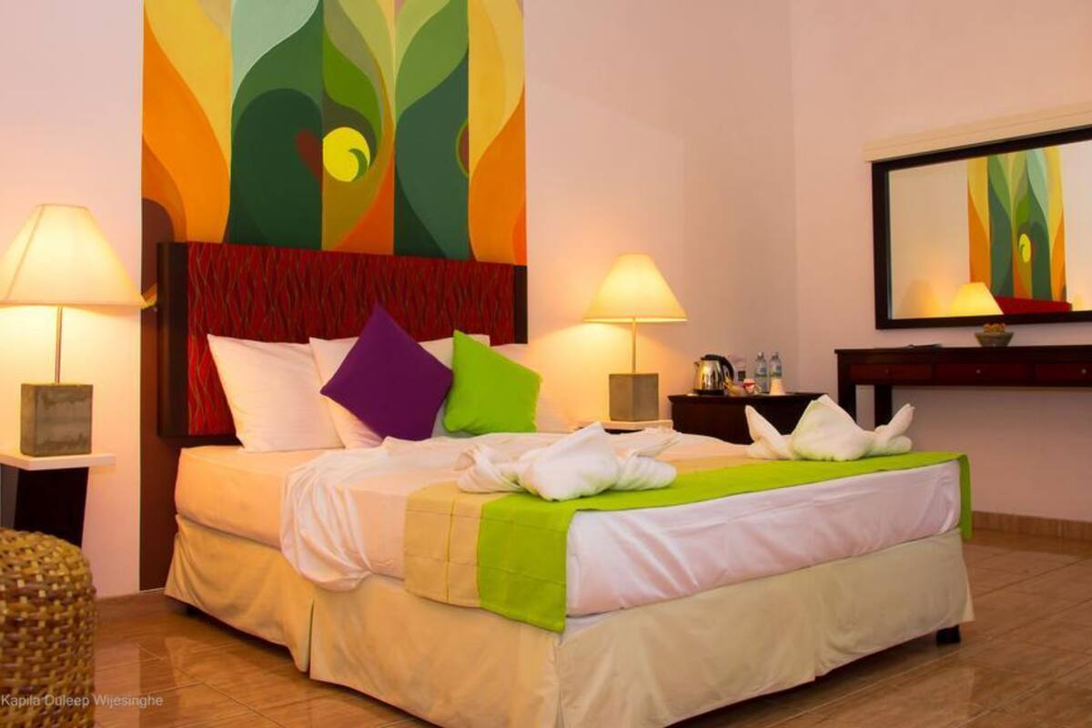 A hotel room with a bed, two bedside lamps, a nightstand, a mirror, and a colorful abstract painting on the wall.